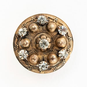 May include: A circular antique brass button with a floral pattern and clear crystal embellishments. The button has a central flower motif, encircled by smaller crystals and raised brass detailing. The button is set against a white backdrop.