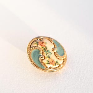 May include: A vintage, round button with a gold-coloured metal frame. The button features a cream-coloured centre with a floral design in pink and green, framed by teal blue accents. The button is likely for sewing or decorative purposes.