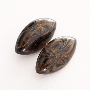 May include: Two marquise-shaped glass beads featuring a dark brown and black swirled pattern with embedded gold-colored glitter accents. The beads have a smooth, polished surface and a pointed, elongated oval shape with a reflective finish.