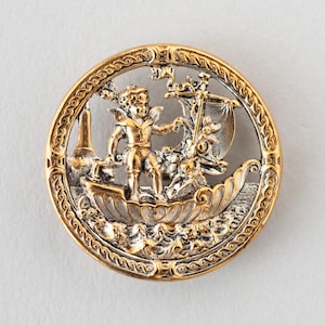 May include: A circular, two-tone metal button with a nautical theme. It depicts a cherub in a boat, with a lighthouse in the background. The outer rim has an ornate, gold-coloured design. The button is likely for decorative purposes.