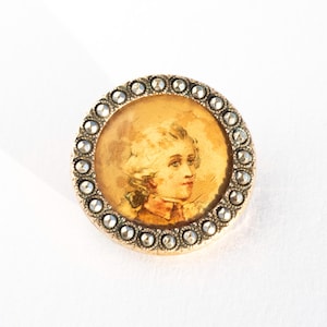 May include: A circular antique brooch featuring a portrait of a man within a gold frame. The portrait is encircled by a ring of small, clear stones. The brooch has a gold-coloured metal base and a pin on the reverse.