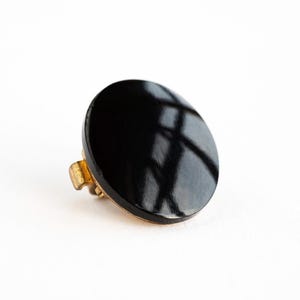 May include: A black, oval ring with a glossy surface. The ring has a gold-coloured setting. The ring is set against a white background.