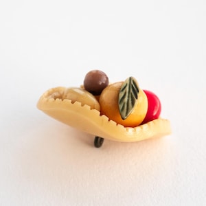 May include: A decorative button shaped like a beige hat with a scalloped brim. The hat is adorned with a small brown sphere, a yellow fruit-like shape with a green leaf detail, and a smooth red sphere resting on the top surface.