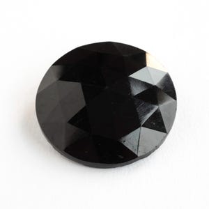 May include: A circular, black button with a faceted surface. The button is made of a shiny, opaque material and has a geometric design. The button is set against a white background.