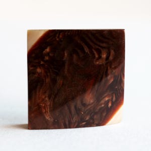 May include: A square, handcrafted soap bar with a marbled design. The soap features swirling patterns of dark brown and tan colors, creating a unique and artistic appearance. The edges are slightly rounded.