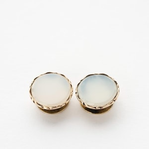 May include: A pair of round, light-colored plugs with a gold-colored, scalloped edge. The plugs have a smooth, opaque surface and a gold-colored base. The jewelry is set against a white background.