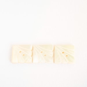 Könnte beinhalten: Drei quadratische, cremeweiße Knöpfe mit Perlmutt-Finish. Jeder Knopf weist ein geschnitztes, fächerartiges Muster auf, das von der Ecke ausgeht, sowie zwei mittige Löcher zur Befestigung. Die Oberfläche hat eine glatte, glänzende Textur.