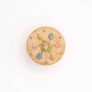 May include: A round, fabric-covered button in a light beige tone, featuring a central green embroidered star-like pattern surrounded by small, alternating yellow, blue, and pink embroidered shapes and tiny green dots across the surface.