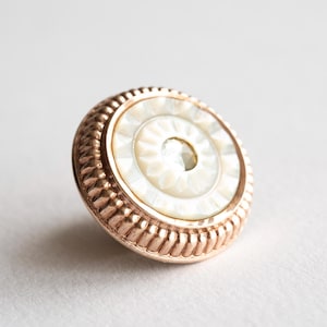 May include: A circular button featuring a central iridescent white mother-of-pearl inlay with a sunburst pattern, surrounded by a metallic rose gold rim with a textured, ribbed edge. The button has a polished finish and a raised, rounded profile.