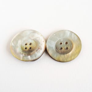 以下が含まれることがあります： 虹色に輝くマザーオブパール素材で作られた2つの丸いボタン。各ボタンの中央はくぼんでおり、縫い付け用の4つの穴が開いています。ボタンの表面は、クリーム色、灰色、薄茶色の色合いが混ざり合った自然な渦巻き状の光沢を見せています。