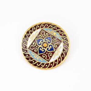 May include: A circular, gold-coloured brooch with an ornate design. The centre has a square pattern with blue, white, and yellow details. The outer ring is dark red with a braided gold border. A decorative accessory.