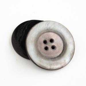 May include: Two large buttons. One is a glossy black, the other is a pearlescent gray with four holes. The gray button has a raised center and a wide, flat rim. The buttons are on a white background.