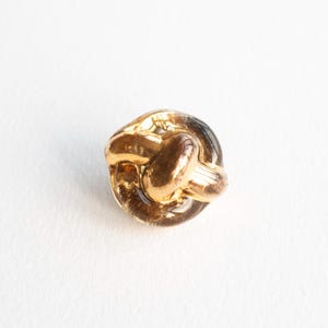Könnte beinhalten: Ein goldfarbener Knopf in Knotenform mit klarer, abgerundeter Basis. Der Knopf hat eine glänzende, metallische Oberfläche und ein detailliertes Design. Der Knopf ist wahrscheinlich für Kleidung oder Bastelprojekte.