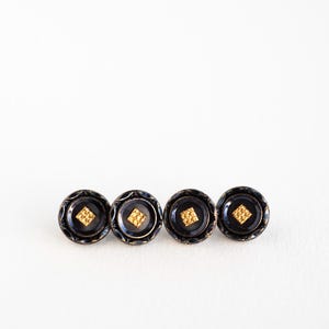 May include: Four black buttons with gold diamond-shaped centres and decorative outer rings. The buttons are arranged in a horizontal line against a white background. These buttons could be used for sewing or crafts.