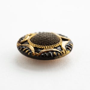May include: A round button featuring a dark brown woven fabric center surrounded by a gold-toned metal border. The outer edge is black with raised gold-toned accents in a repeating pattern, creating a textured and layered appearance.