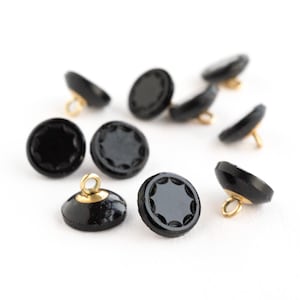 Könnte beinhalten: Eine Sammlung schwarzer, runder Knöpfe mit einem dekorativen Sternmuster auf der Vorderseite. Jeder Knopf hat eine goldfarbene Metallschlaufe auf der Rückseite zum Annähen. Die Knöpfe sind aus glänzendem Material.