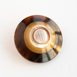 May include: A round button with concentric circles of varying colors. The outer ring is a dark brown, followed by a gold band, and a central iridescent white circle. The button has a glossy finish.