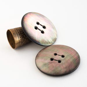 May include: Two iridescent, oval buttons with four holes each, and a brass thimble. The buttons have a pearlescent sheen with shades of grey, green, and pink. The thimble is a warm gold colour with a textured pattern.
