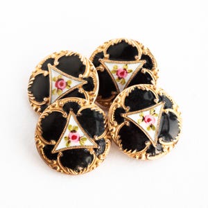 May include: Four round buttons featuring a black enamel center with a gold filigree border. Each button has a central white triangular inlay decorated with a small pink rose and green leaves. The gold metal edges are textured with ornate detailing.