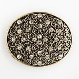 May include: Oval-shaped, antique gold-tone metal brooch with a filigree design. The brooch features a pattern of swirling metalwork and is studded with numerous clear, round rhinestones. The border is a lighter gold color.