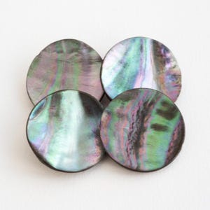 May include: Four circular buttons crafted from iridescent abalone shell, featuring a smooth, polished surface with swirling patterns of deep purple, vibrant green, and soft blue hues that shift in colour depending on the light reflection.