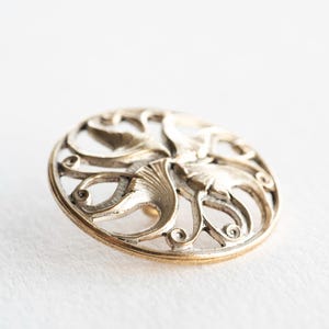 May include: A circular metal button featuring an intricate openwork design with raised, swirling leaf-like patterns. The button has a metallic gold and silver-toned finish with darkened recessed areas that highlight the detailed filigree texture.