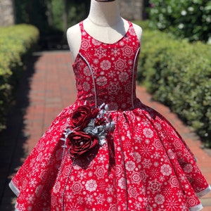 Puede incluir: Un vestido de estampado de copo de nieve rojo y plateado con ribete plateado y un adorno de rosa roja. El vestido tiene una falda completa y un corpiño sin mangas.