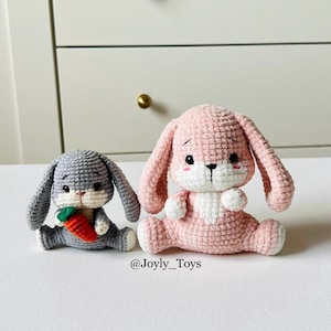 以下が含まれることがあります： 2つの編みぐるみウサギのおもちゃ。1つはグレーで、小さなオレンジ色のニンジンを持っています。もう1つはピンクです。どちらも長い耳と白いアクセントがあり、座っています。テキスト「@Joyly_Toys」が下にあります。