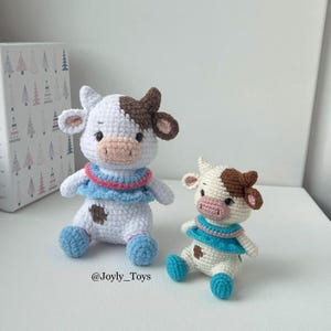 以下が含まれることがあります： 茶色と白の毛並みの、編みぐるみのかわいい牛のぬいぐるみが2体。1体はもう1体より大きく、どちらも青とピンクのドレスを着ています。大きい方の牛は足を組んで座っており、小さい方の牛は足を広げて座っています。2体とも黒い目とピンクの鼻をしています。