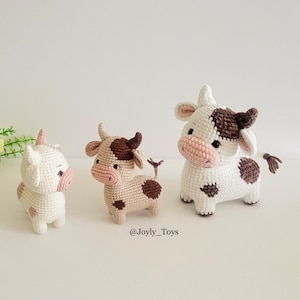 May include: Three crocheted cow plushies. The plushies are white with brown spots and pink noses. The plushies are standing on a white surface. The text '@Joyly_Toys' is below the plushies.