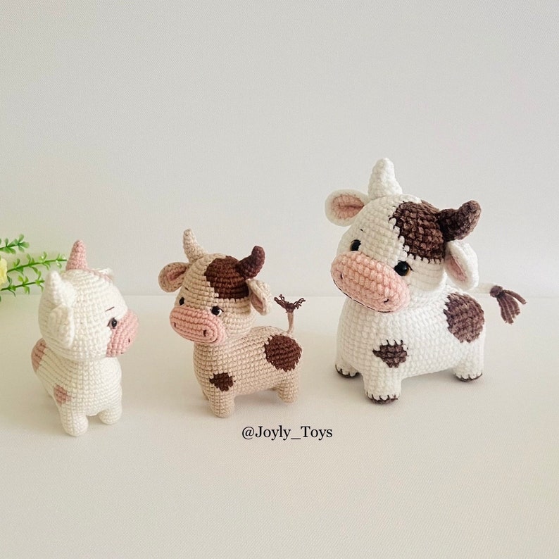 May include: Three crocheted cow plushies. The plushies are white with brown spots and pink noses. The plushies are standing on a white surface. The text '@Joyly_Toys' is below the plushies.