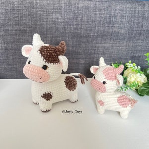 May include: Two crocheted cow plushies. The larger cow is white with brown spots and a pink nose. The smaller cow is white with pink spots and a pink nose. Both cows have black eyes. The larger cow is standing on a white surface and the smaller cow is standing on a white surface with a green plant in the background. The text '@Joyly_Toys' is visible on the white surface.