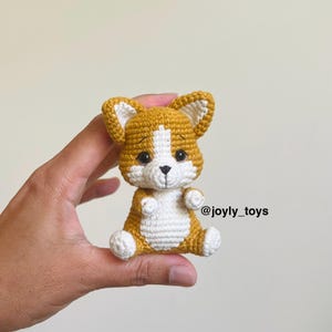Può includere: Un piccolo giocattolo corgi lavorato all'uncinetto a mano con filato marrone dorato e bianco. Il peluche ha orecchie a punta, occhi scuri e un naso ricamato. Il testo @joyly_toys è visibile sul lato destro del giocattolo su uno sfondo neutro.