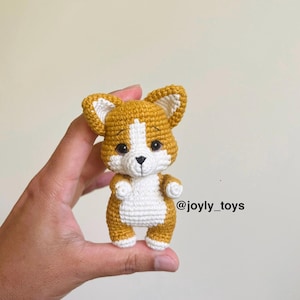 Puede incluir: Un pequeño perro amigurumi de ganchillo en hilo dorado y blanco. La figura presenta orejas puntiagudas, ojos de seguridad oscuros, una nariz bordada en negro y una mancha blanca en el pecho. El texto @joyly_toys aparece impreso en negro.