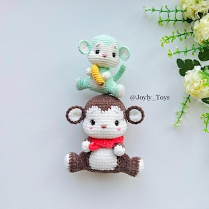 Può includere: Due scimmie all'uncinetto fatte a mano. La scimmia inferiore è marrone e bianca con una sciarpa rossa. La scimmia superiore è verde chiaro e tiene in mano una banana gialla. I giocattoli sono su una superficie bianca.