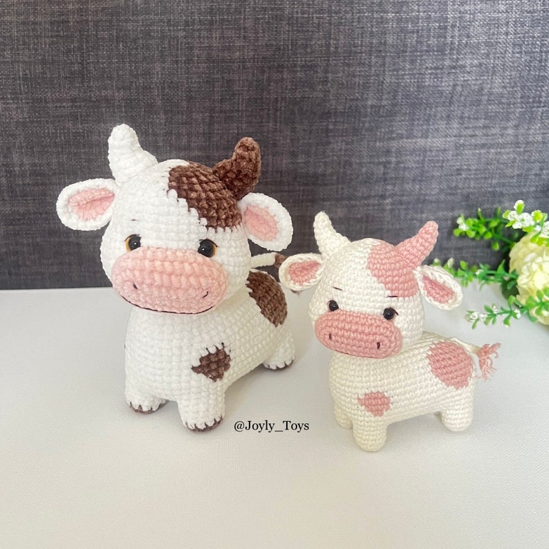 May include: Two crocheted cow plushies. The larger cow is white with brown spots and the smaller cow is white with pink spots. Both cows have pink noses and black eyes. The smaller cow is standing to the right of the larger cow. The text '@Joyly_Toys' is visible below the cows.