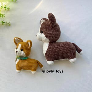 Può includere: Due giocattoli a forma di cane realizzati all'uncinetto. Il più piccolo è marrone dorato e bianco con un collare verde. Il più grande è marrone scuro e bianco. Entrambi hanno orecchie a punta e occhi neri. Il testo @joyly_toys è stampato in nero.