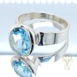 Może przedstawiać: Srebrny pierścionek z jasnoniebieskim owalnym kamieniem szlachetnym. Pierścionek jest osadzony w oprawie typu bezel i ma polerowane wykończenie.
