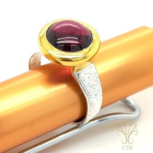 Könnte beinhalten: Ein silberner Ring mit einem goldenen Band und einem runden, dunkelvioletten Edelstein.