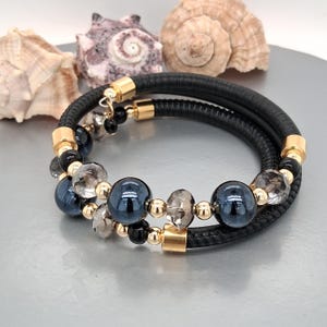 May include: Black leather wrap bracelet with gold-tone accents and beads. The bracelet features dark blue and clear faceted beads, along with smaller black beads. The bracelet is displayed on a silver surface with seashells in the background.