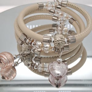 May include: A stack of cream-colored bracelets with silver-toned accents and clear beads. Each bracelet has a spiral design. The bracelets are adorned with heart-shaped and round pink glass charms. The bracelets are displayed on a reflective surface.