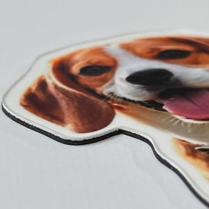 May include: A die-cut magnet featuring a realistic, high-resolution color photograph of a beagle dog's face. The dog has brown and white fur, dark eyes, and an open mouth with a visible pink tongue. The magnet has a thick black edge.