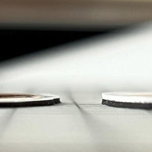 May include: Two thin, flat, circular objects are positioned side-by-side. The object on the left features a dark brown top surface with a white border, while the object on the right has a white top surface. Both have a black base layer.