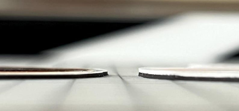 May include: Two thin, flat, circular objects are positioned side-by-side. The object on the left features a dark brown top surface with a white border, while the object on the right has a white top surface. Both have a black base layer.