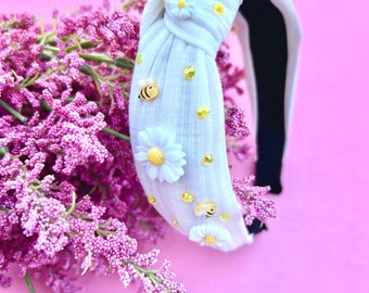 May include: White knotted headband with a black elastic band. The headband is decorated with white daisies, yellow bees, and gold glitter.