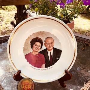 Puede incluir: Un plato de cerámica blanco con un borde dorado presenta un retrato del presidente y la señora Lyndon B. Johnson. El plato está sobre un soporte de madera con un pequeño botón "For the USA".