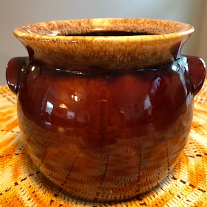 以下が含まれることがあります： 光沢のある茶色の陶器のポットで、幅広のタン色の縁と両側のハンドルが付いています。 ポットは丸みを帯びた形で、黄と白のかぎ針編みの表面に置かれています。 ポットの内部は暗いです。