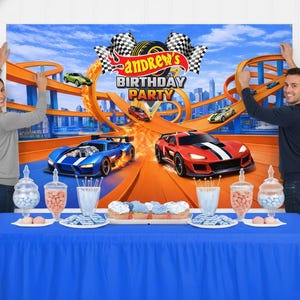 May include: A rectangular birthday party backdrop featuring a race car theme with orange tracks, a blue car, a red car, and checkered flags. The text reads Andrew's Birthday Party. The backdrop is displayed behind a blue table with candy jars.