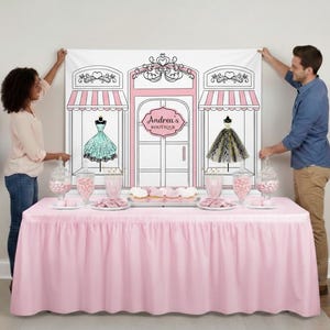 May include: A rectangular backdrop featuring a boutique storefront illustration with pink striped awnings, two dress mannequins, and the text Andrea's Boutique, displayed above a table covered with a pink cloth and filled with glass candy jars.
