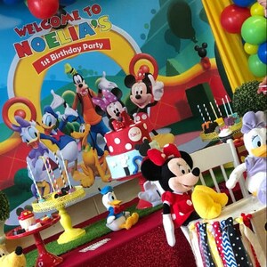 Puede incluir: Una escena de fiesta de cumplea&ntilde;os con Mickey Mouse y sus amigos. Una tarta con lunares rojos y un letrero que dice "Noelia's 1st Birthday Party" es el centro. Globos y decoraciones en rojo, amarillo y azul completan el ambiente alegre.